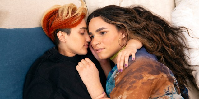A transmasculine gender-nonconforming person with short blond and bright red hair and a transfeminine non-binary person with long brown wavy hair, waking up together in bed. The person with brown hair is looking into the distance as though thinking about something.