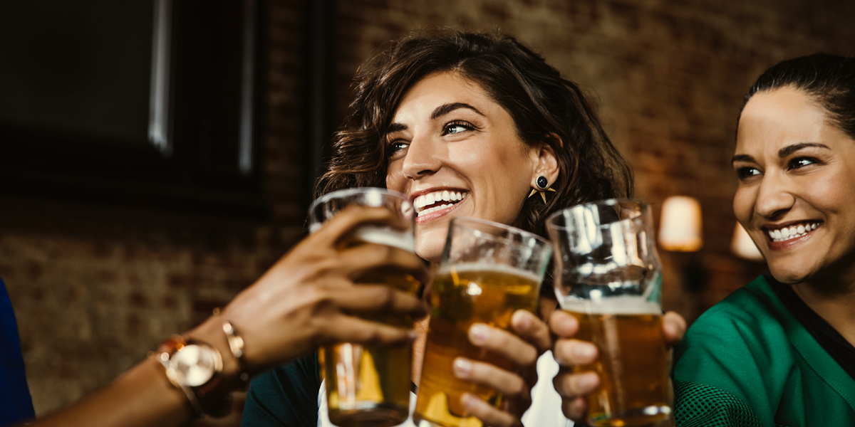 A group of three women drink beer and smile together, the woman in the center has brown curly hair in a bob. They are in front of a brick wall.