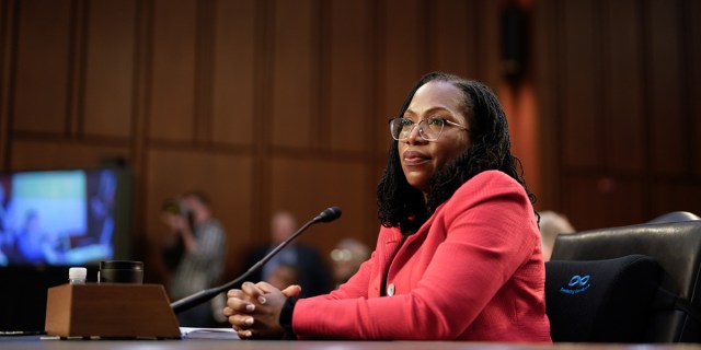 Ketanji Brown Jackson, a Black woman with dark skin, round glass, and shoulder length dreadlocks, is in a red blazer as she sits alone at a desk in front of Congress.