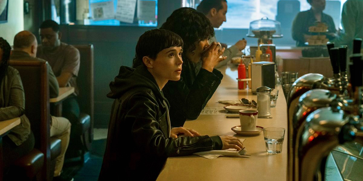 Elliot Page as Viktor Hargreeves in a still from The Umbrella Academy. Victor is in a leather jacket and is ordering coffee at a diner. The photo appears to be shot at nighttime, with lots of shadows.