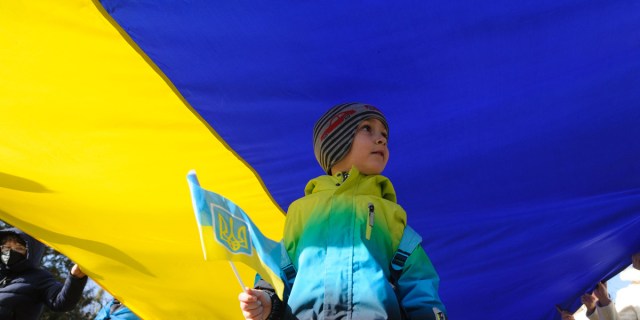 A young boy stands underneath the Ukranian flag while also holding a small Ukrainian flag
