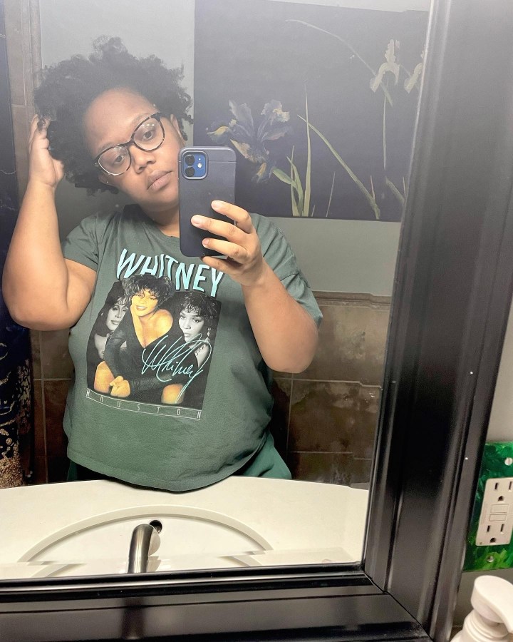 Carmen, a Black woman with mid-length curly hair is wearing a Whitney Houston tee shirt