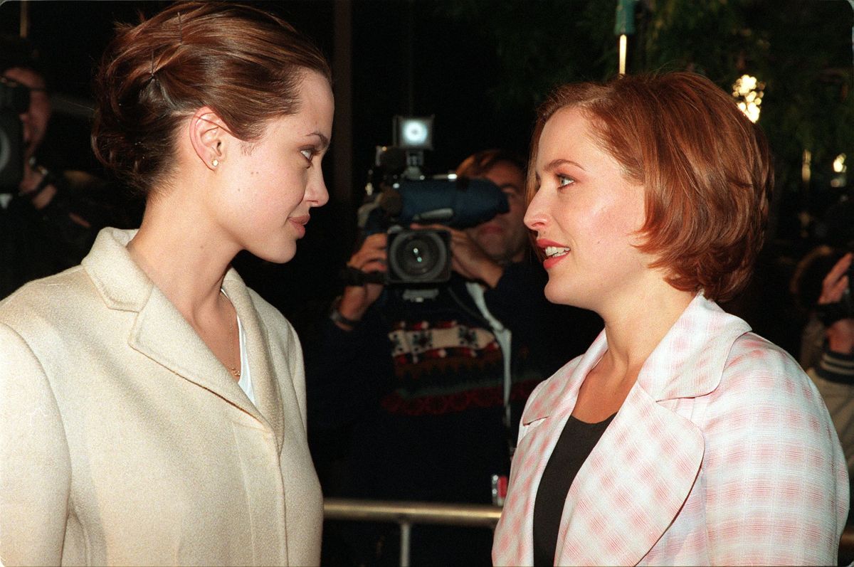 Angelina Jolie is in a white dress blazer that is closed with her brunette hair swept into a bun, she's talking with Gillian Anderson, with red hair in a bob and a checkered suit coat.