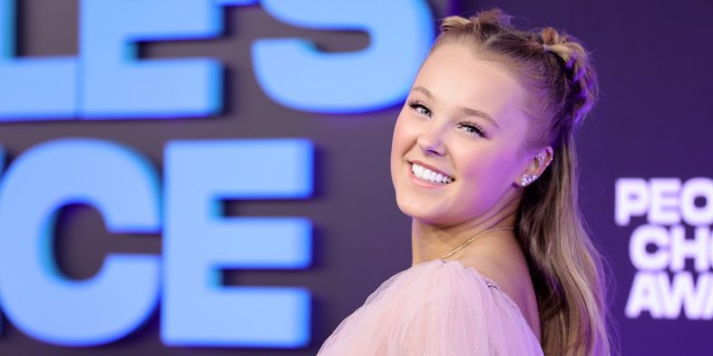 JoJo Siwa smiles brightly in a close up on the red carpet, her hair is pulled back and down and she has on a flowy pink top