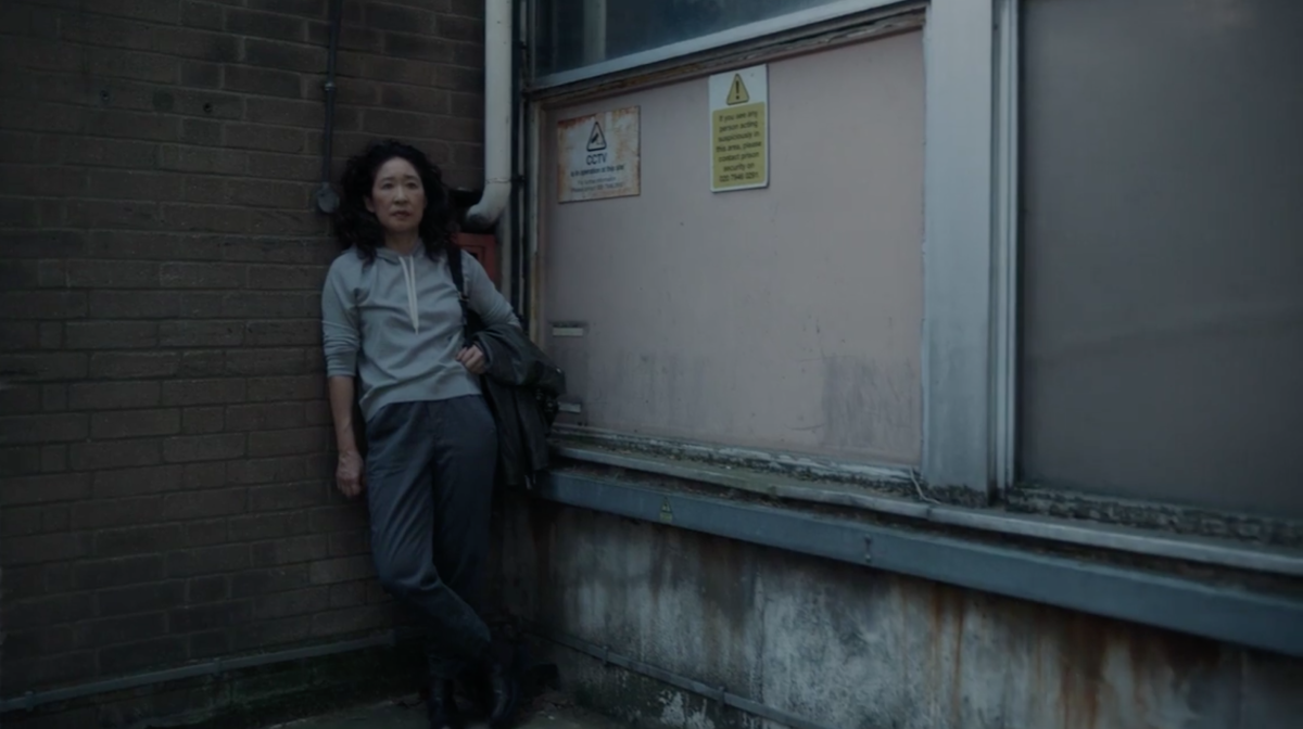 Eve (Sandra Oh) wears a gray hoodie and gray slacks while leaning against a brick wall on Killing Eve