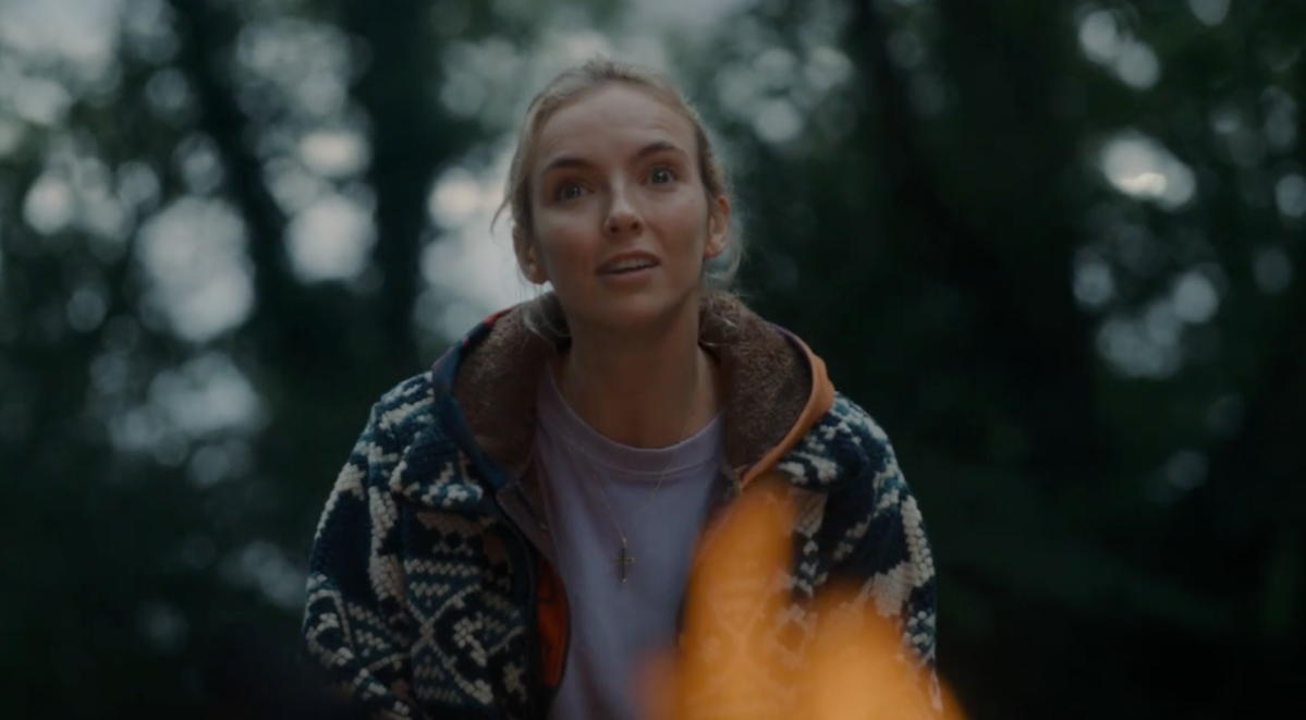 Villanelle (Jodie Comer) wears a fluffy jacket and a pale purple t-shirt and sits behind a fire