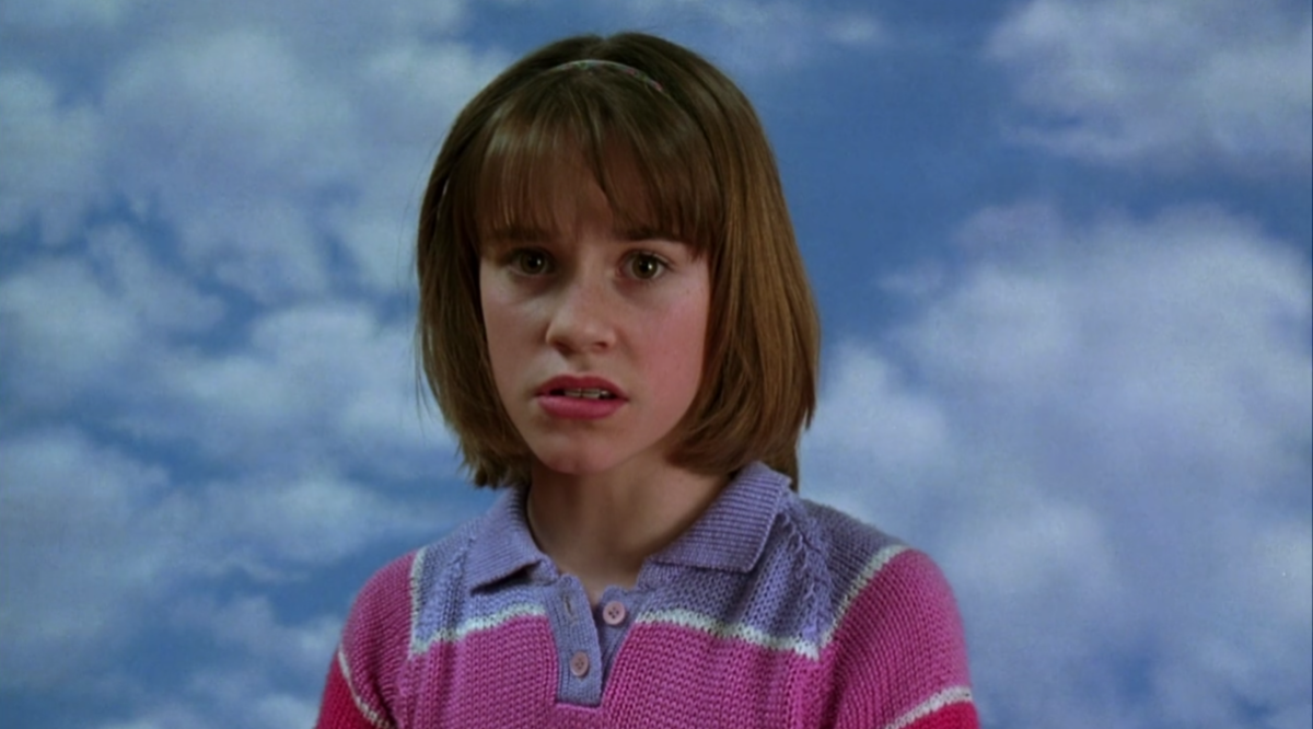 A young Jenna Rink has a bob haircut and bangs and a mouthguard. She is wearing a collared sweater with purple, pink, and white stripes. She is posing for school picture day.