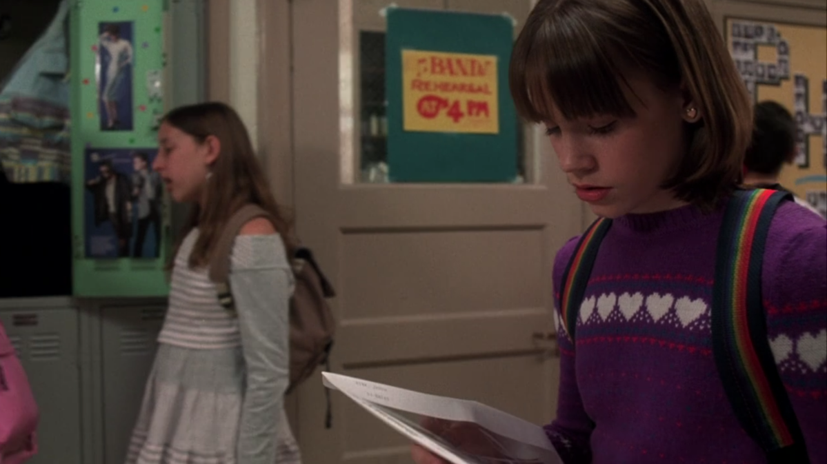 A young Jenna Rink has a short bob and bangs and wears a purple sweater with white hearts on it. Her backpack has rainbow straps.