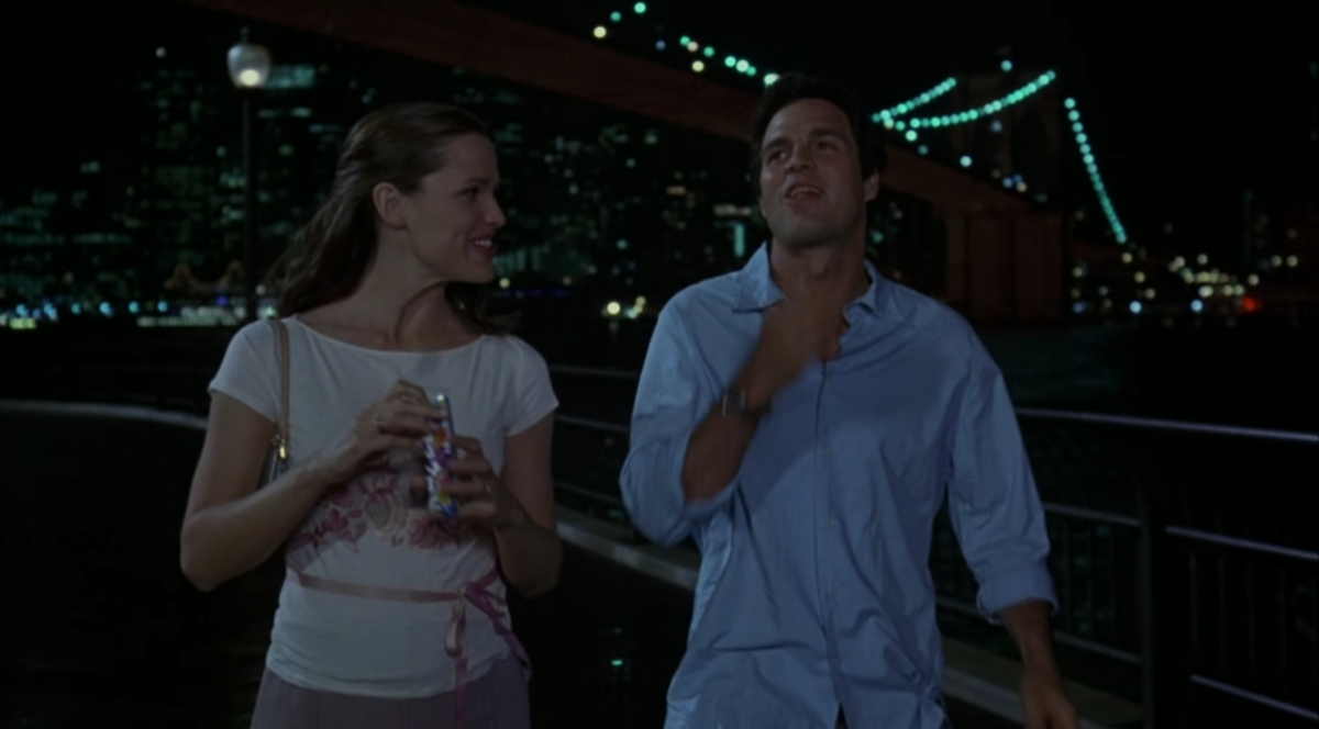 Jenna and Matt are walking with the NYC skyline behind them, eating Razzles. Jenna is wearing a t-shirt with a pink ribbon tied around her middle.