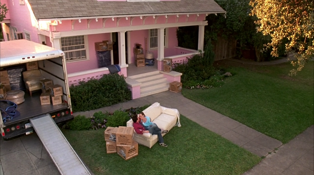 A packed U-Haul sits in front of a pink house. On the lawn, there is a white couch, and Matt and Jenna are snuggling on it.