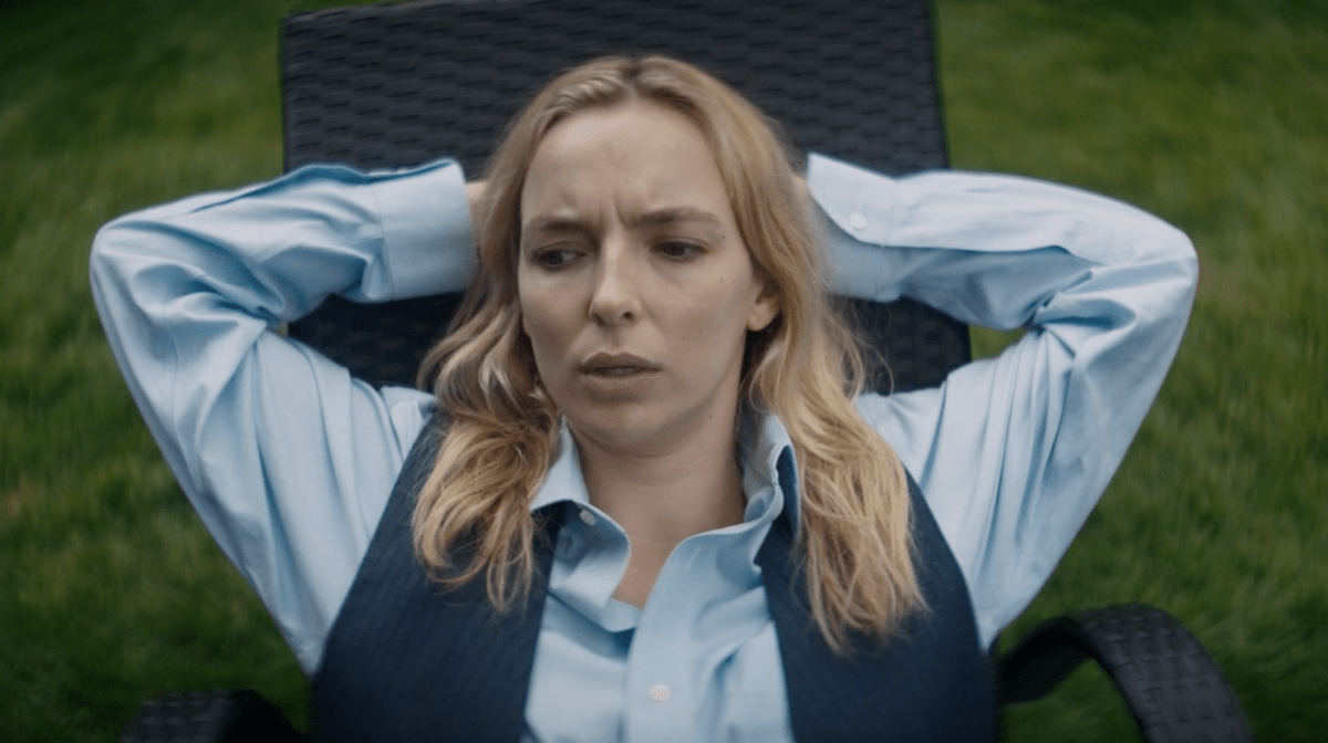 Villanelle (Jodie Comer) is wearing a blue buttondown and a gray vest and has her arms behind her head while she reclines in a lawn chair on Killing Eve.