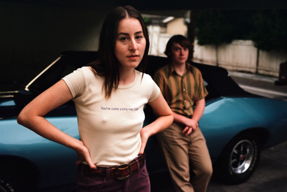 Alana haim and Cooper Hoffman in "Licorice Pizza", Cooper leaning on a blue car, Alana with her hands on her hips wearing a "you've come a long way baby" tiny tee