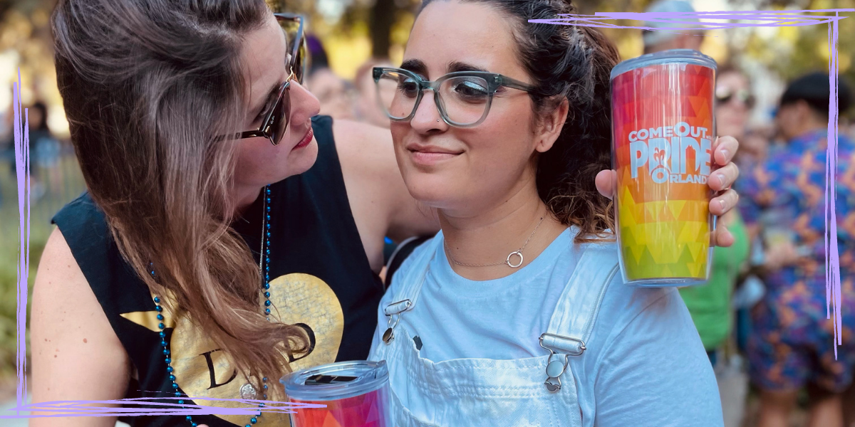 Kristen Arnett, a white queer person in sunglasses and a tanktop that says Dad, leans toward her girlfriend, Autostraddle Managing Editor Kayla Kumari Upadhyaya, who is a South Asian and white lesbian with her brown curly hair in a ponytail and wearing glue glasses and white overalls over a white t-shirt. They are at Orlando Pride, and Kristen is holding a cup that says Orlando Pride Party on it.