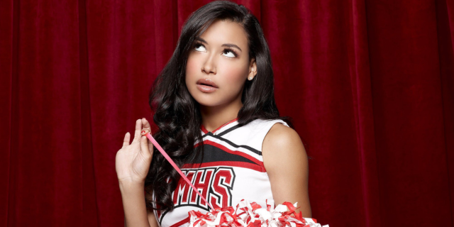Santana Lopez (Naya Rivera) from Glee wears a cheerleading uniform while rolling her eyes and pulling at a string of her pom-pom