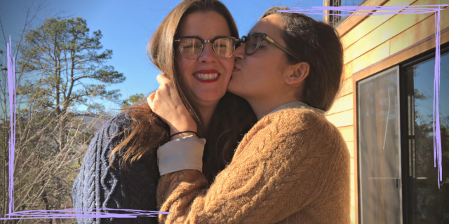 This is a photo of Kayla and Kristen. Kayla embraces Kristen and kisses them on the cheek while Kristen smiles. They both have long brown hair and wear glasses. They're also both wearing sweaters!