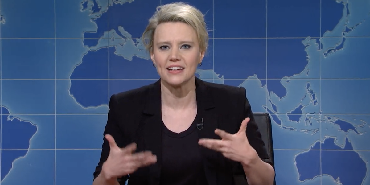 Kate McKinnon in a black blazer gesticulating with her hands in front of the blue globe Weekend Update desk on SNL
