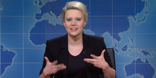 Kate McKinnon in a black blazer gesticulating with her hands in front of the blue globe Weekend Update desk on SNL