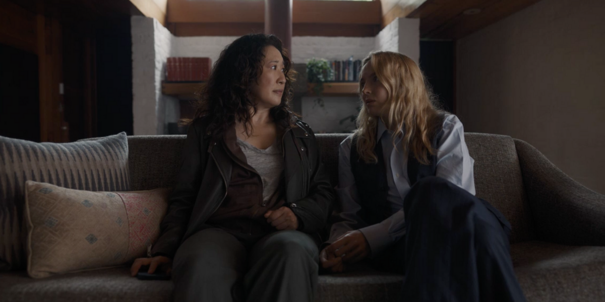 Eve (Sandra Oh) sits next to Villanelle (Jodie Comer) on a couch. Eve looks serious. Villanelle leans toward her and is wearing a suit.
