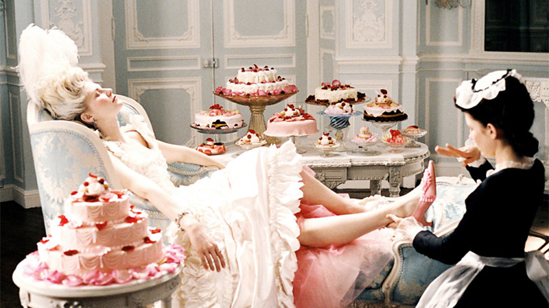 Marie Antoinette sits with her feet up in front of a table of cakes