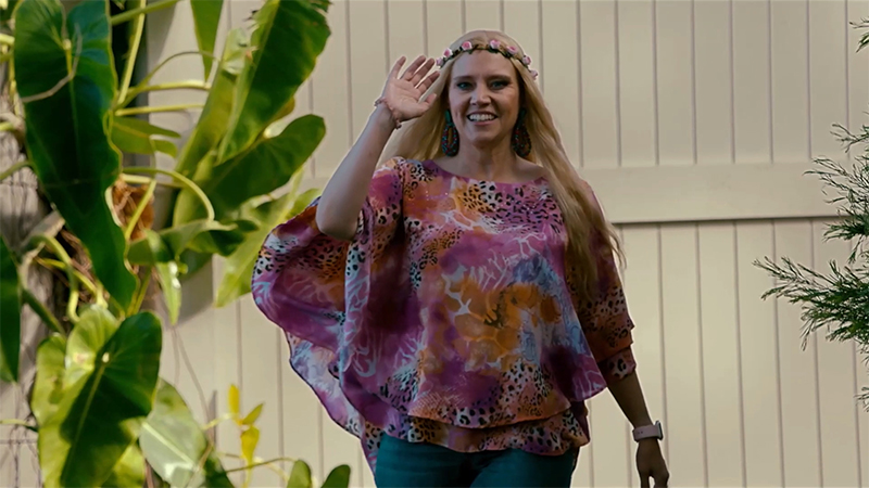 Kate McKinnon as Carole Baskin in a pink leopard print shirt, waving