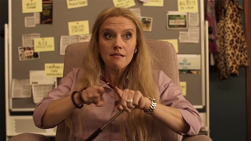 Kate McKinnon as Carole Baskin in a pink shirt sitting at her desk
