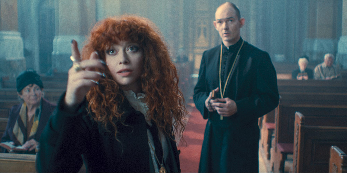 Natasha Lyonne in a black suit pointing, with a priest looking on in the background