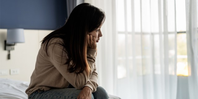 A woman with long, brown hair wearing jeans and a beige sweater sits on the edge of a bed with a white bedspread. She rests her chin on her hand. In the background, there are open windows covered by gauzy white curtains.