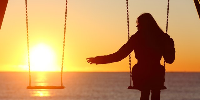 Single woman in a swing on the beach at sunset reaches out to the empty swing beside her, seems sad