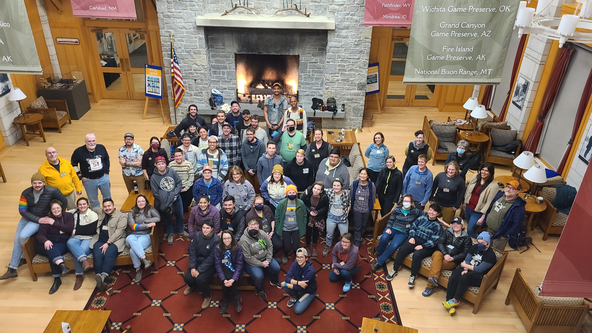 Shot from above, a collection of roughly 30 attendees in front of a lit fire place in a open room with wood floors.