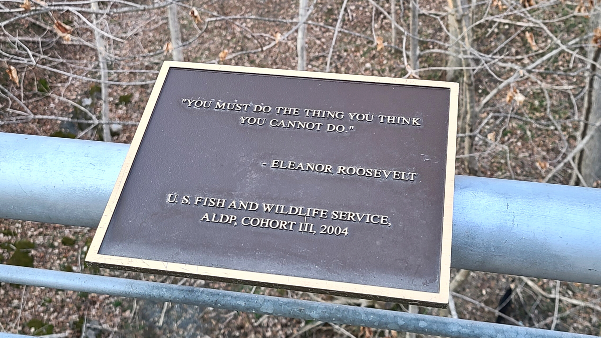 A close up of a bronze plaque on a metal railing. The plaque reads: "You must do the thing you think you cannot do" by Eleanor Roosevelt, from the U.S. Fish and Wildlife Service, ALDP, Cohort Ill, 2004. Behind the plaque are bare trees.