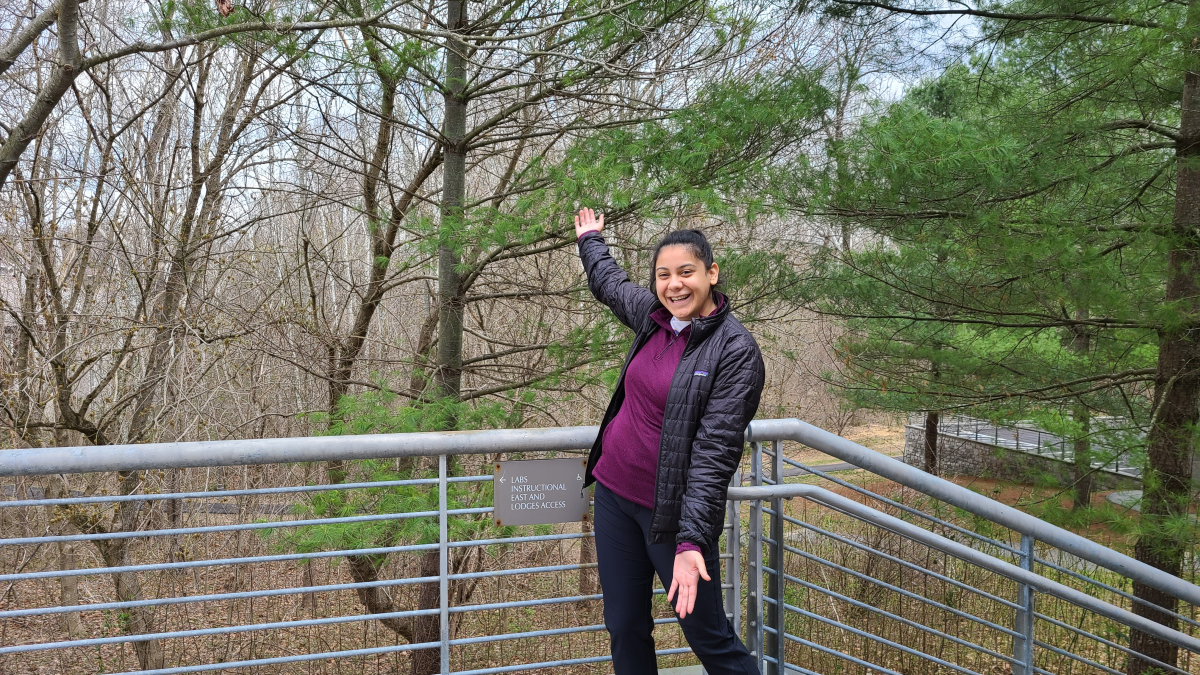 Jessica is a Latina woman of Mexican heritage who wears her hair in a ponytail. She is smiling and pointing to the green trees while standing on a patio. She is wearing a purple shirt with a black light weight puffer coat over it.