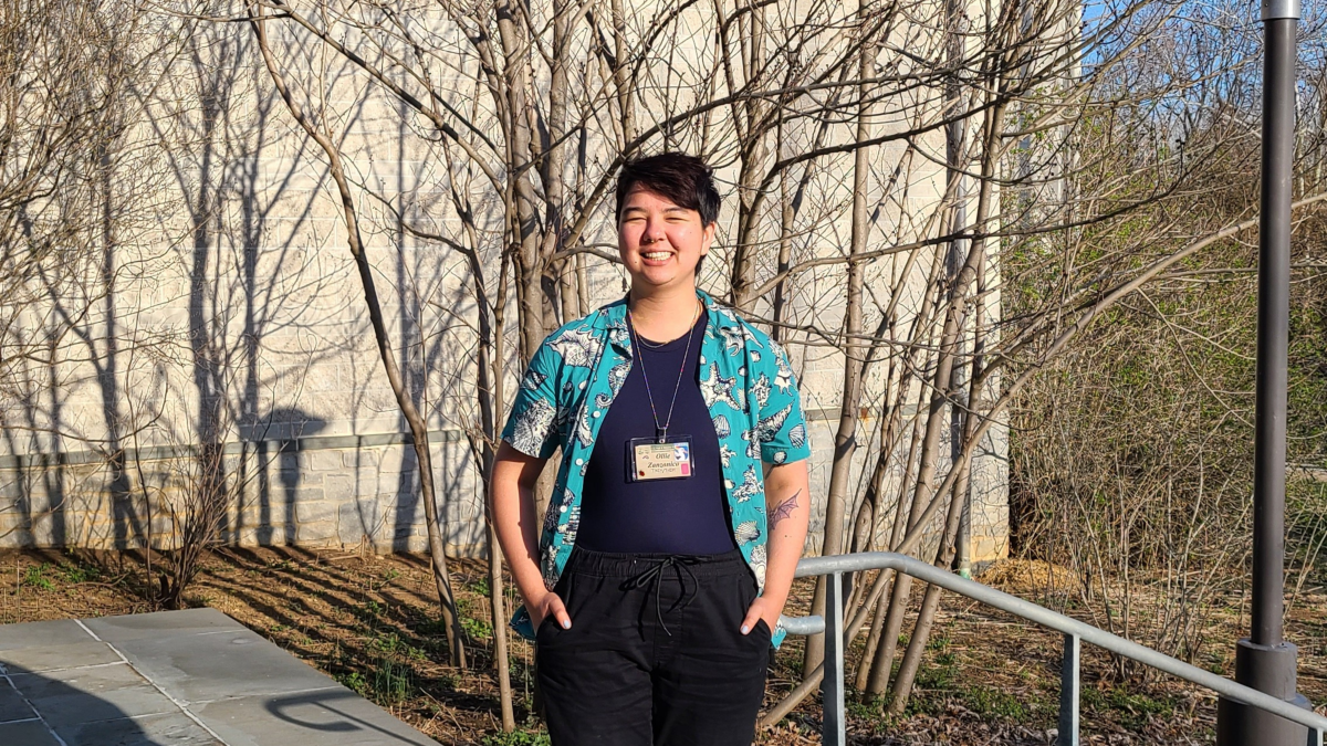 Ollie is a person of color with short dark hair that frames over their right eye. They are smiling brightly on a sunny day in front of bare trees. They have on a short sleeved aqua blue top with a pattern on it, which is layered over navy blue top. They are wearing a name badge.