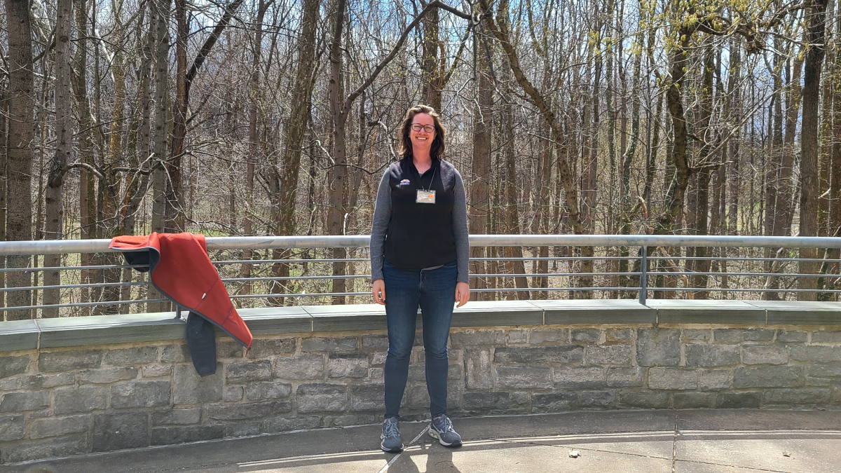 Hannah is a white cis woman in dark blue jeans and a grey and black baseball sleeved shirt. She has shoulder length brown hair and glasses and is wearing sneakers. She stands in front of a stone half wall. The half wall is on a ledge overlooking a forest of bare trees.