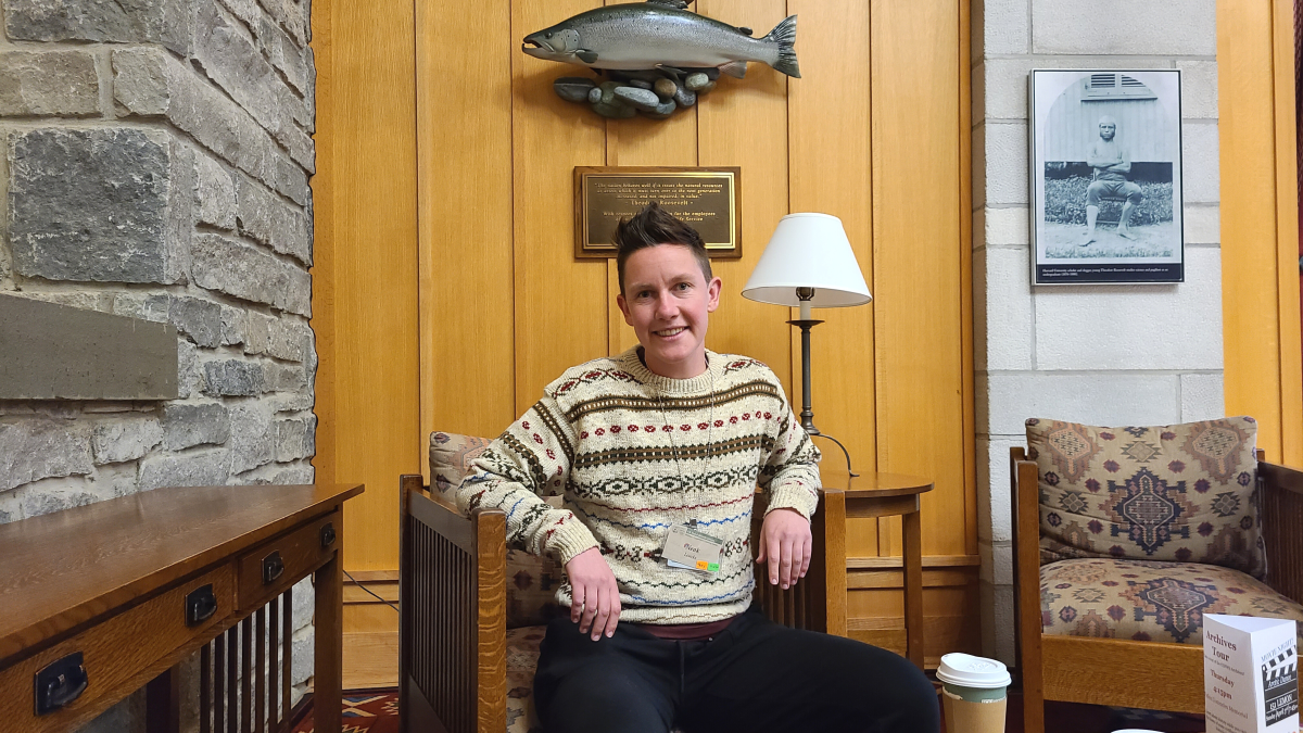 Neak Loucks is a white person with short brunette hair and an undercut. They are wearing a warm beige sweater with green and red designs. They are sitting in a wood chair in front of a wood paneled wall with a fish hangin got it and a stone side. They are smiling.