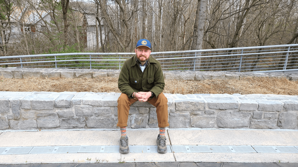 Luke Warner is a white a trans man in a green jacket with pockets and caramel brown trousers. He is wearing a blue baseball cap and has a brunette beard. He is sitting on a stone halfway in front of a bare forest.