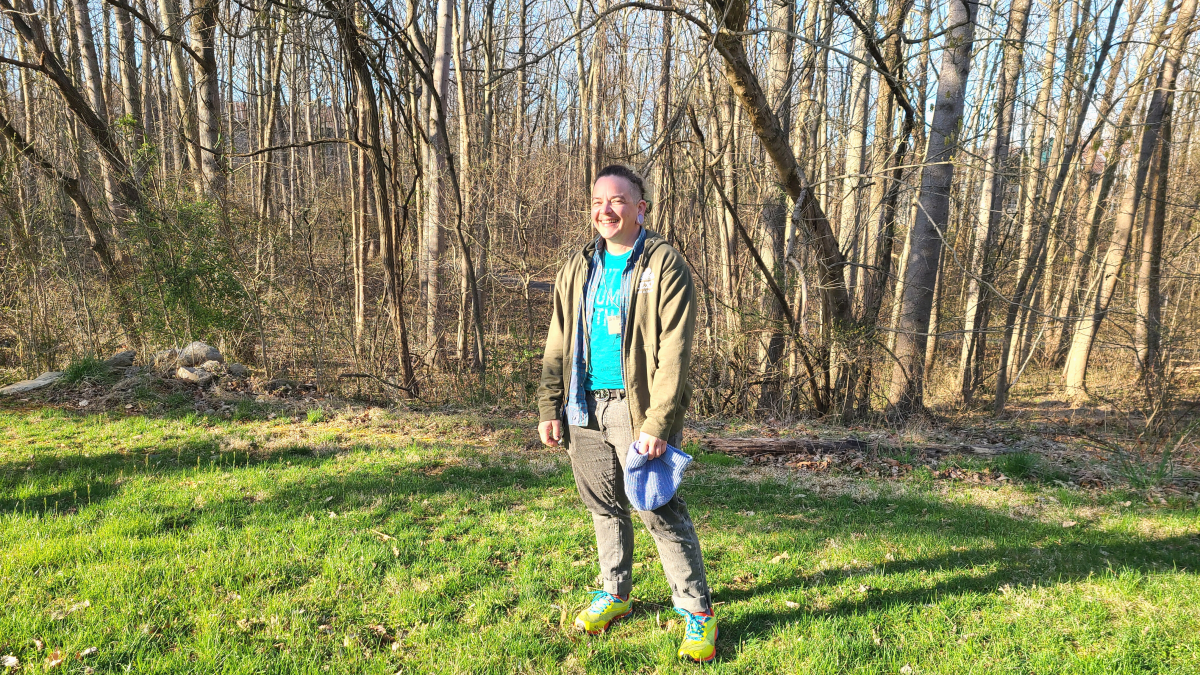 Scout is a person of color with short cut brunette hair, an aqua blue shirt, a olive green jacket, and khaki pants. They are holding a blue beanie in their hand. They are standing and smiling brightly to the left of camera. Scout stands on bright green grass in front of a forest of bare trees.