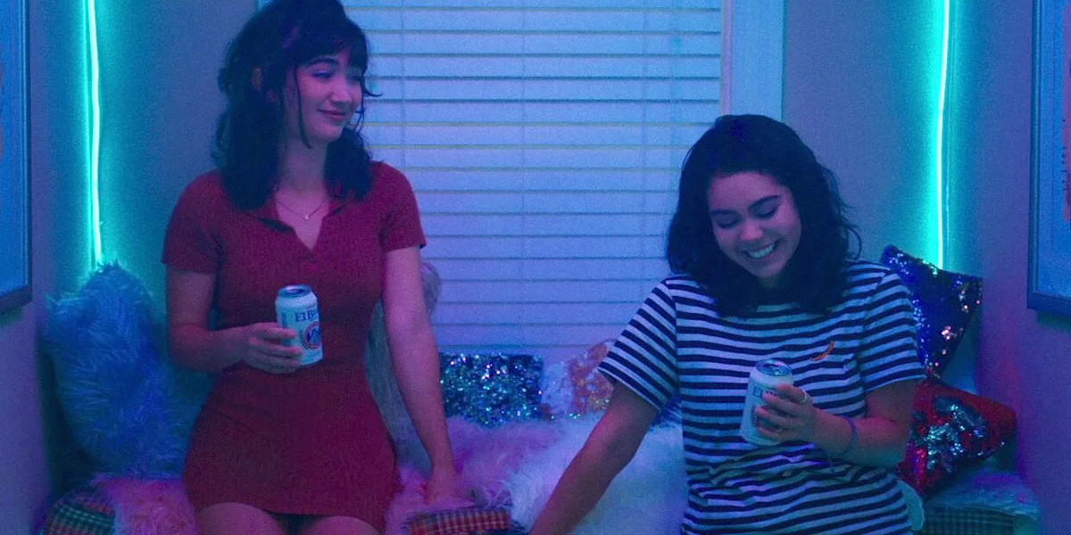 In a still from Hulu's crush, Rowan Blanchard's Paige is smiling while facing right of camera, she is making Auli'i Cravalho's AJ blush. The lighting is shades of blue and purple. Paige is in a red dress with brunette hair half up in a pony tail. AJ is in a black and white striped shirt with brunette wavy hair in a messy bob.