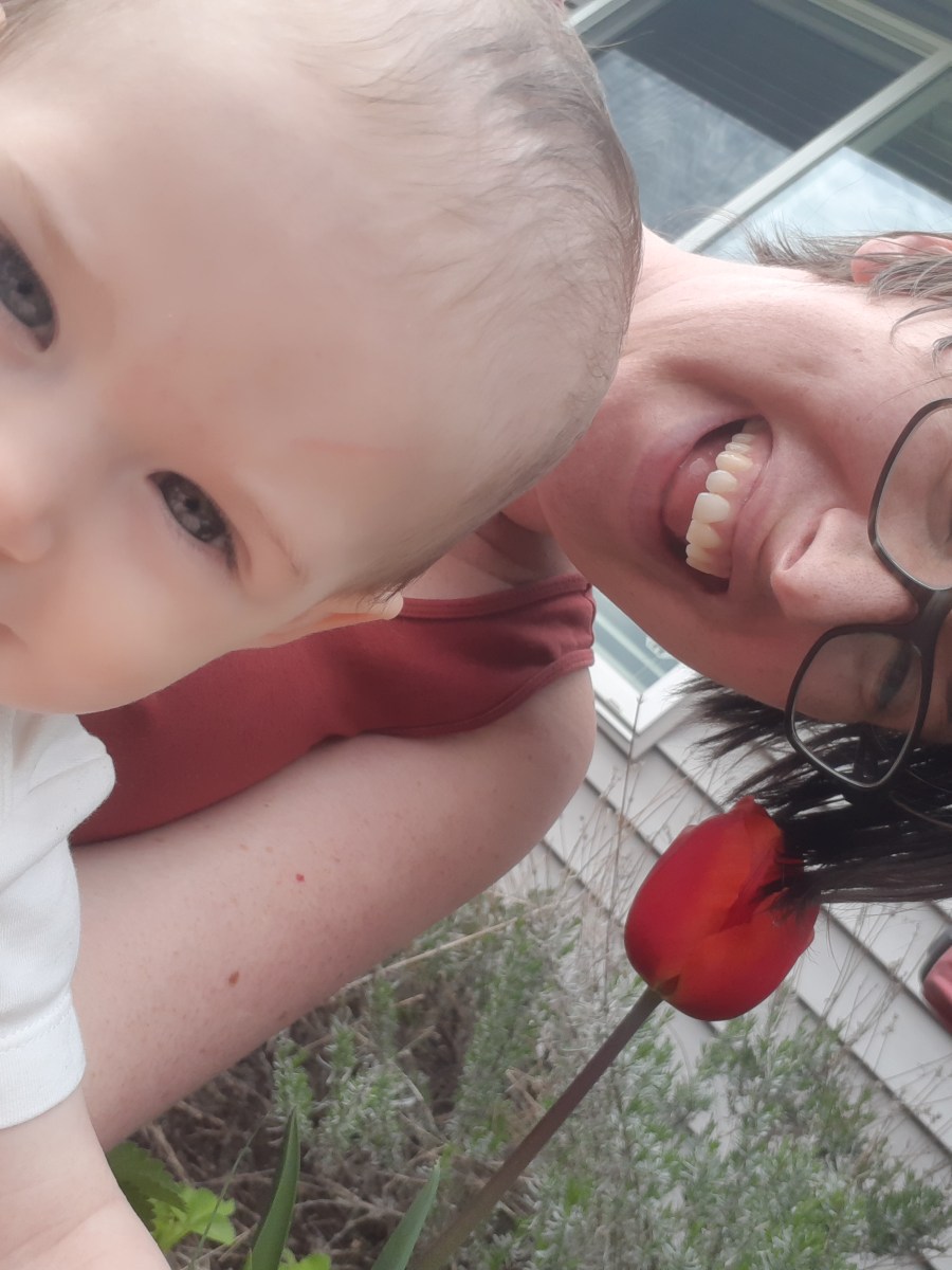 Casey with her baby! Casey is a white woman with short brown hair and glasses. They are laughing in front of a single red tulip.