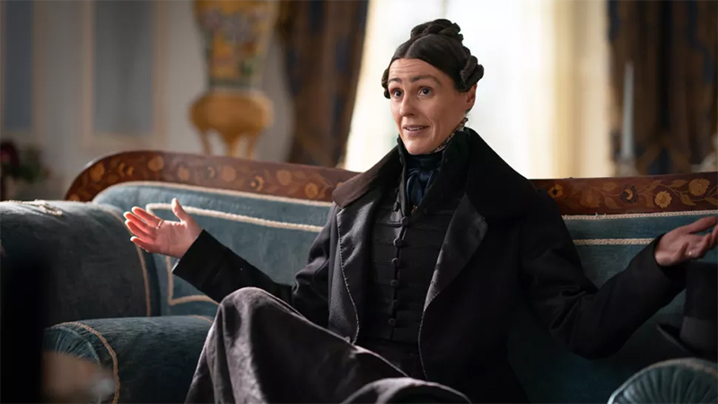 Anne Lister sits on a couch with her arms outstreched