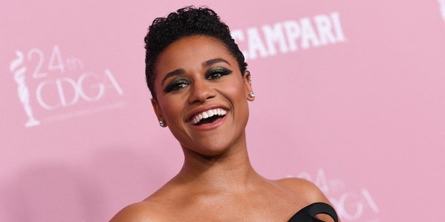 Ariana DeBose smiling in a black dress in front of a pink background