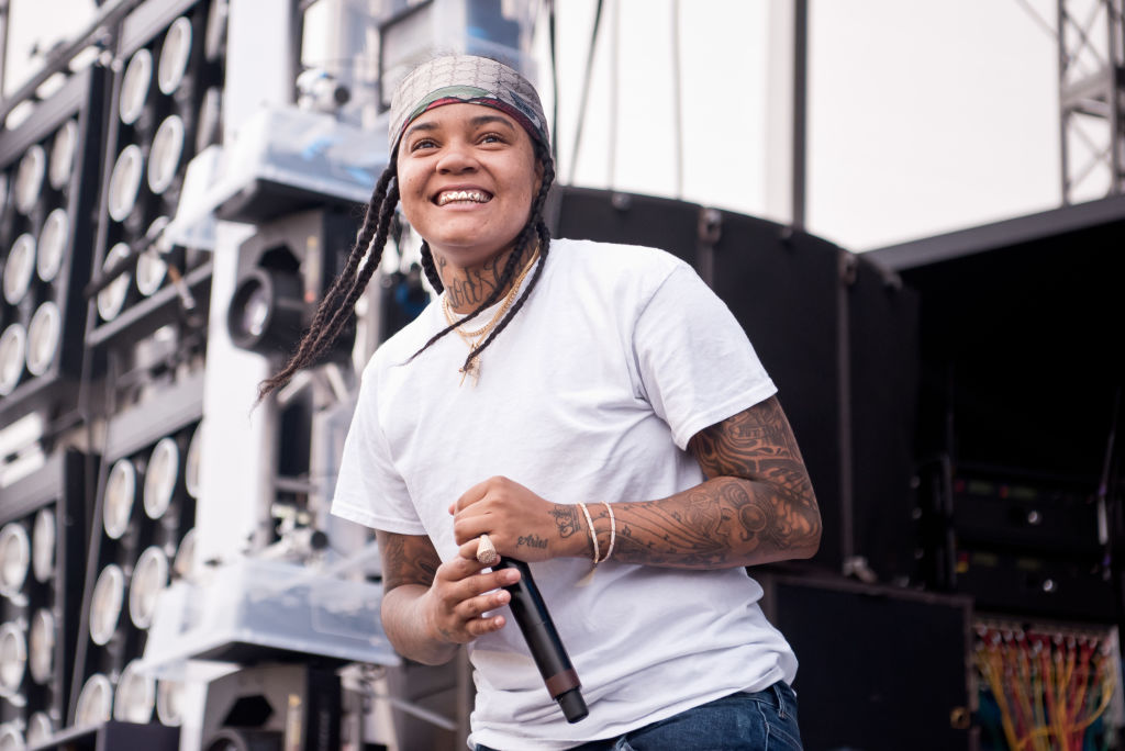 Young M.A holds a microphone and wears a white t-shirt while smiling