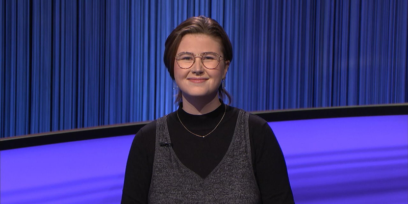 Mattea Roach is on Jeopardy in a grey sweater dress and black turtleneck. She has glasses and brunette hair in a low ponytail. The stage behind her is Navy Blue. She is smiling.