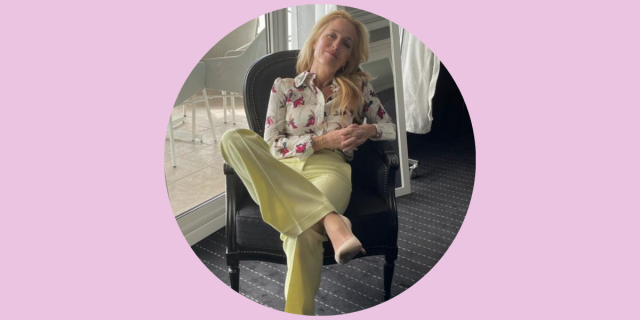 Gillian Anderson sits in a chair with her legs crossed and her arms folder. She's wearing green pants and a floral print buttondown shirt.