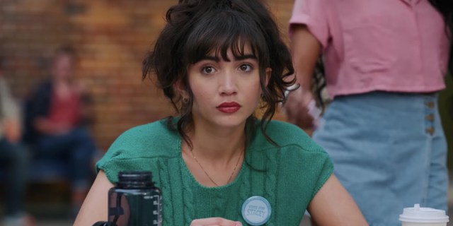 In a still from the movie Crush, Rowan Blanchard as Paige has on red lipstick and short sleeve turquoise sweater, she is sitting at a high school lunch table with her hair in a messy updo.