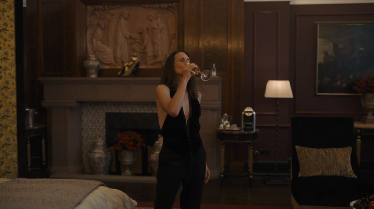 Helene wears a black vest with no shirt underneath over black pants while drinking a flute of champagne on Killing Eve