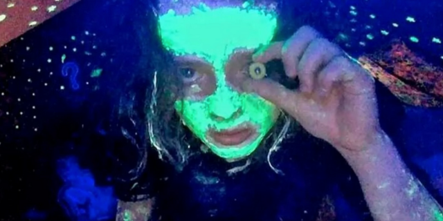 Casey, played by Anna Cobb, looks into her webcam with paint on her face that shines green in her blacklight. She holds a fake eye in front of her left eye in Jane Schoenbrun's movie We're All Going to the World's Fair.