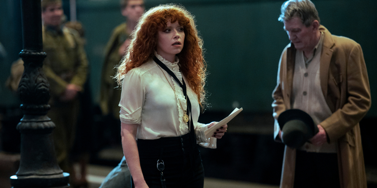 Natasha Lyonne as Nadia stands on a train platform wearing a white button down blouse with a black tie accent in Russian Doll season two.