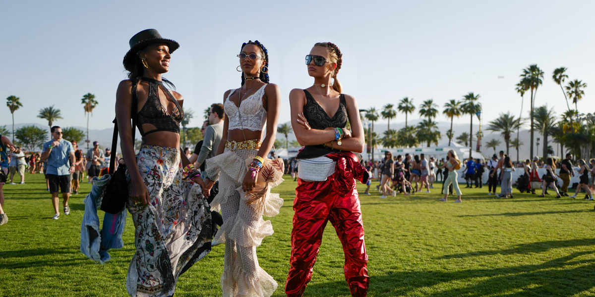Three women in festival closing (the one on the left wears a cowboy hat, the one in the middle has shimmery pants, and the one on the right is in sunglasses) are standing together on the Coachella lawn