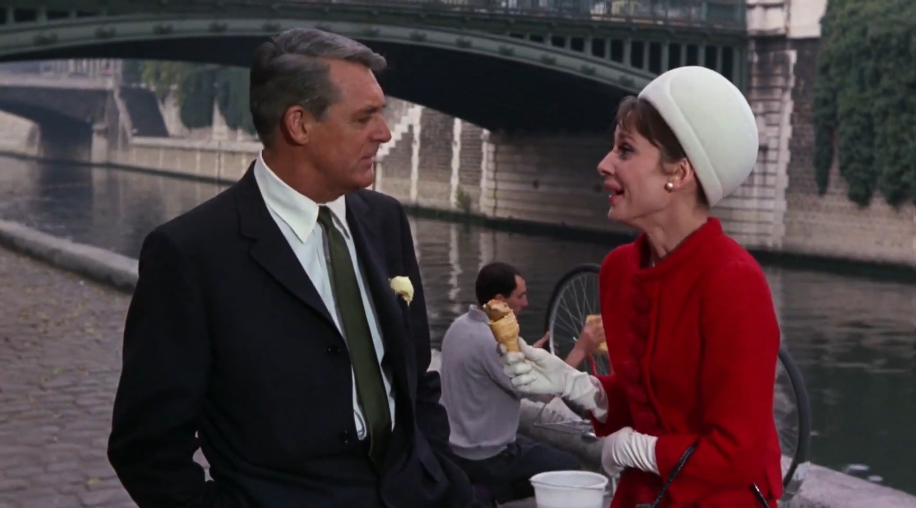 In the movie Charade, Cary Grant and Audrey Hepburn stand near water and a bridge. Cary is wearing a black suit and tie, and Audrey is wearing a red dress and a white hat and white gloves. She holds an ice cream cone.
