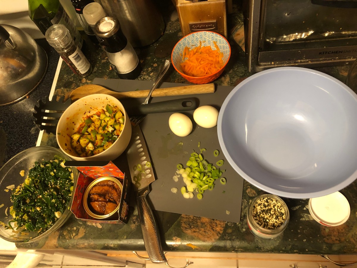 A bowl of spinach, a bowl of smashed cucumbers, a bowl of pickled carrots, chopped scallions, eggs, and a tin of fish are scattered on a countertop with a knife and large mixing bowl